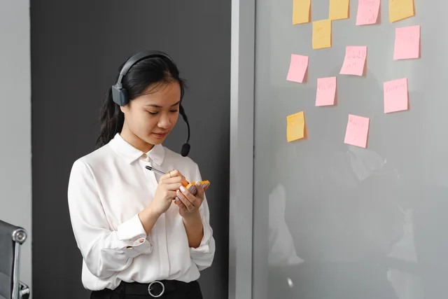 A woman in a call center writing on sticky notes with a headset indoors. Kosten einer wiederkehrenden Servicepanne sichtbar machen