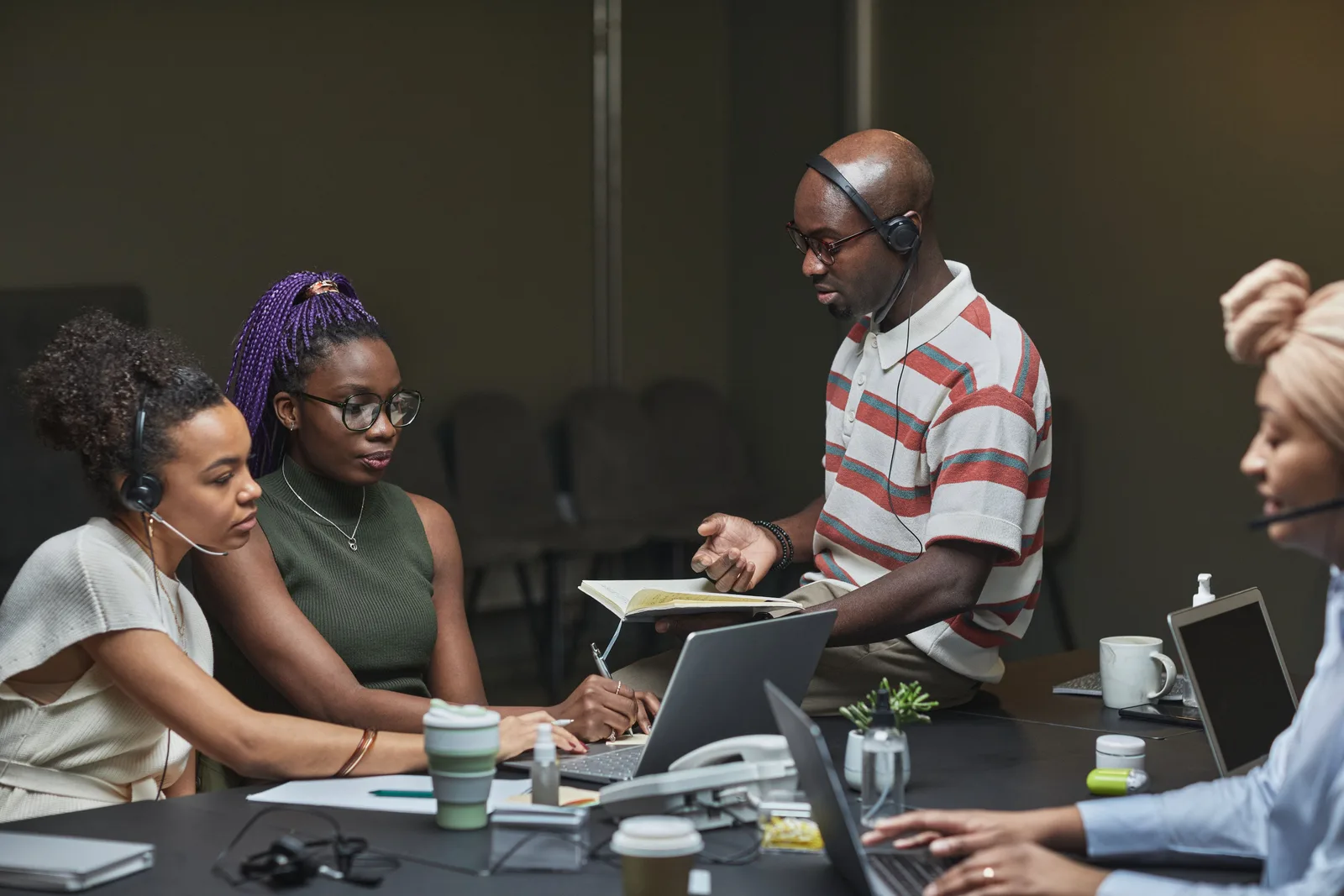 A diverse group of professionals collaborating at a desk in a modern office with laptops and headsets.
