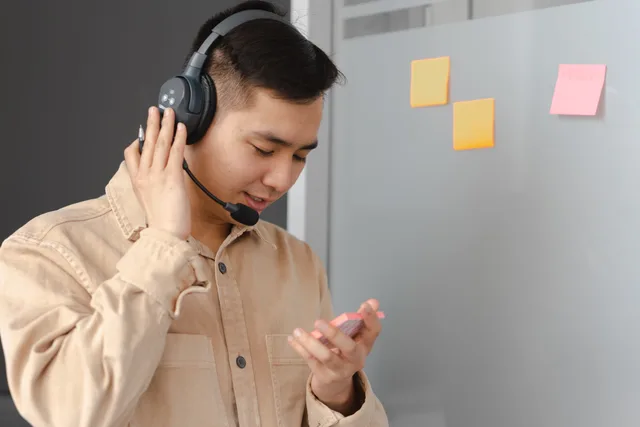 Asian man wearing headset, consulting with clients, holding notes in front of sticky notes. Lebenszyklus-Kommunikation